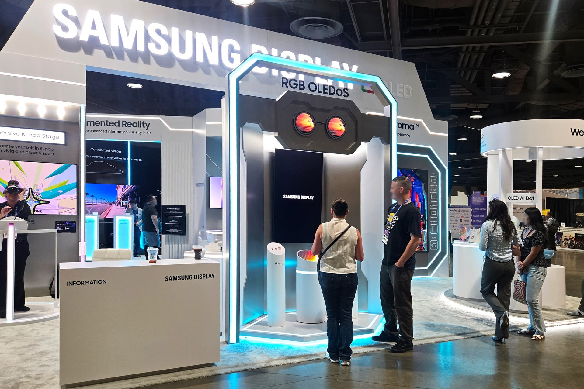People explore Samsung Display exhibits at a tech convention. Bright lights illuminate screens and Samsung XR displays, with visitors interacting and viewing information at various booths. The atmosphere is modern and high-tech.