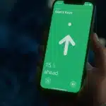 A hand holds a smartphone displaying a green screen with an arrow pointing up, text reading "Alan's Keys," and "15 ft ahead," showing distance to the keys using Apple Find My accessories.