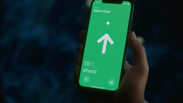 A hand holds a smartphone displaying a green screen with an arrow pointing up, text reading "Alan's Keys," and "15 ft ahead," showing distance to the keys using Apple Find My accessories.