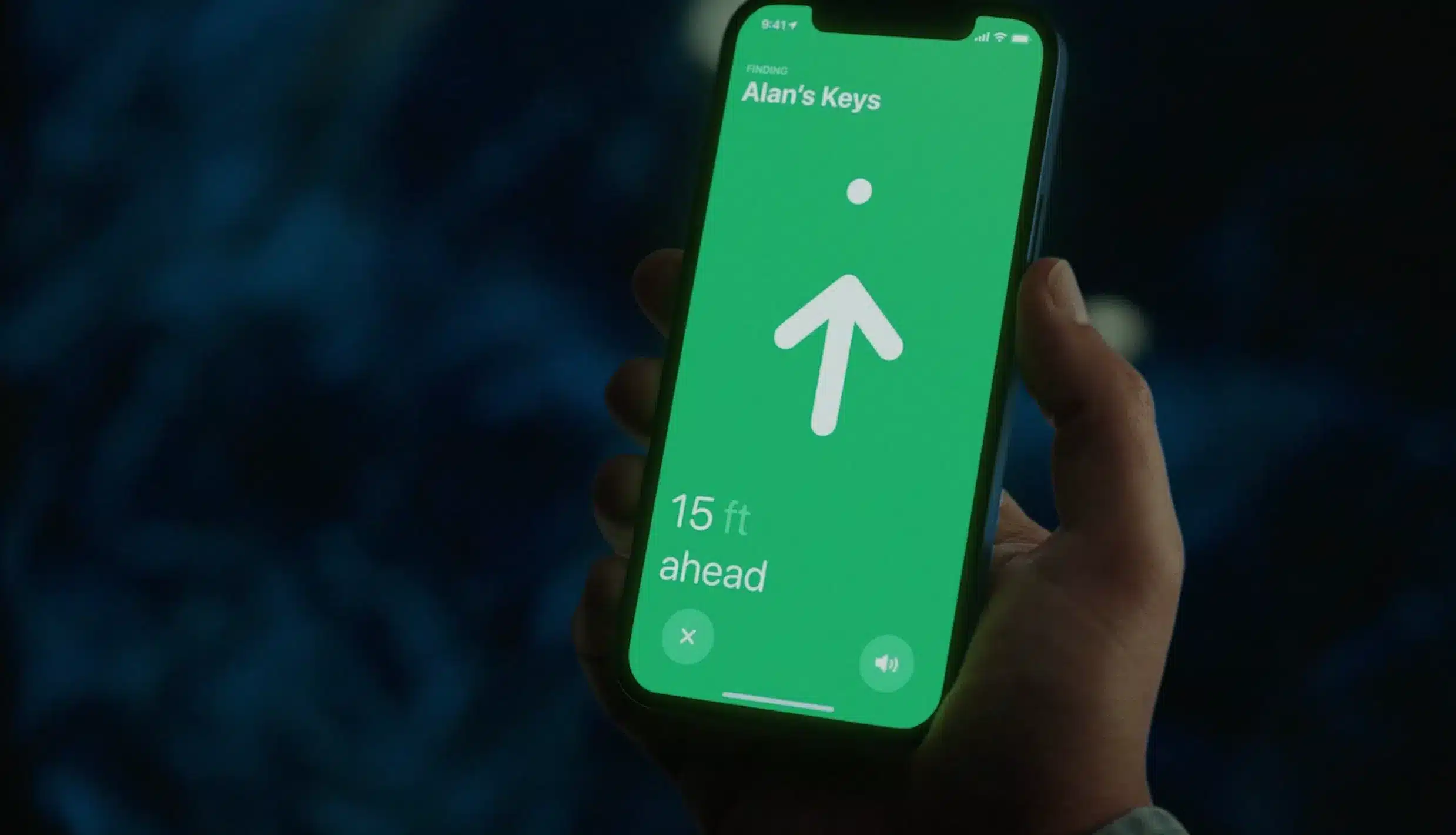 A hand holds a smartphone displaying a green screen with an arrow pointing up, text reading "Alan's Keys," and "15 ft ahead," showing distance to the keys using Apple Find My accessories.