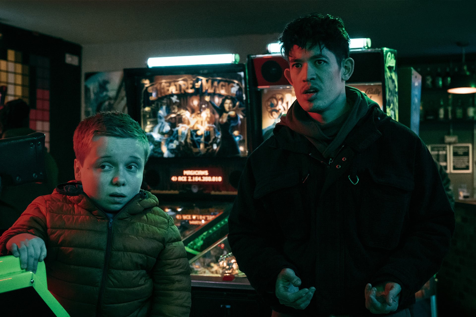 A young boy in a brown puffer jacket and an older man reminiscent of Gary Oldman in Slow Horses stand before illuminated pinball machines in an arcade, both looking surprised or concerned, as if caught in a suspenseful Apple TV moment.