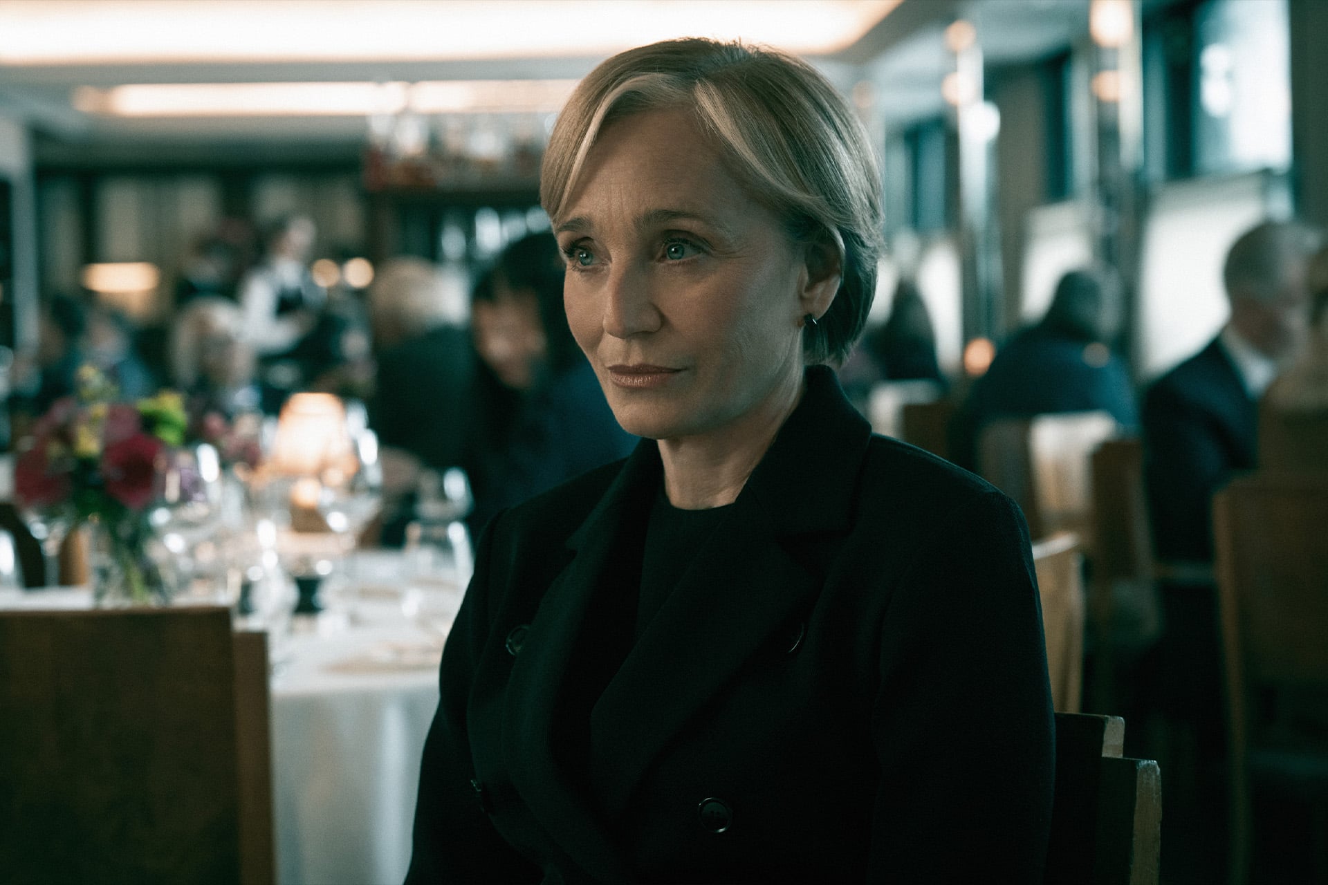 A woman with short blonde hair, dressed in a black coat, sits at a table in an upscale, softly lit restaurant—evoking the moody intrigue of Mick Herron's Slow Horses season 6—with other people dining in the background.