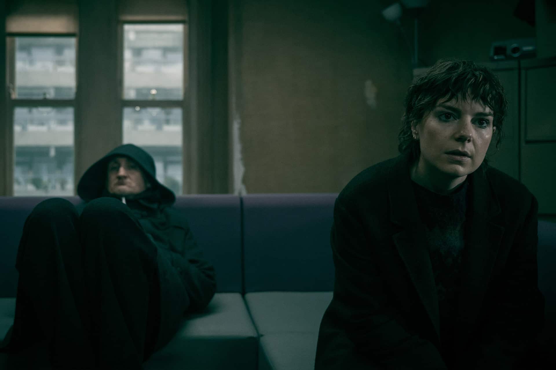 Two people sit on a couch in a dimly lit room. One looks concerned, while the other sits further back in a hood, knees up. Light filters through windows behind them—a moody setting reminiscent of Gary Oldman’s Jackson Lamb in Slow Horses season 6.