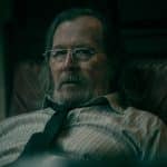 A middle-aged man with long gray hair, glasses, and a beard sits in a leather chair, wearing a striped shirt and a loosely knotted tie, evoking Gary Oldman's serious look from Slow Horses in the moody dim lighting.