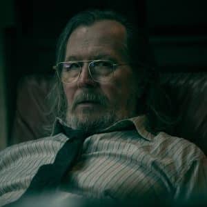 A middle-aged man with long gray hair, glasses, and a beard sits in a leather chair, wearing a striped shirt and a loosely knotted tie, evoking Gary Oldman's serious look from Slow Horses in the moody dim lighting.