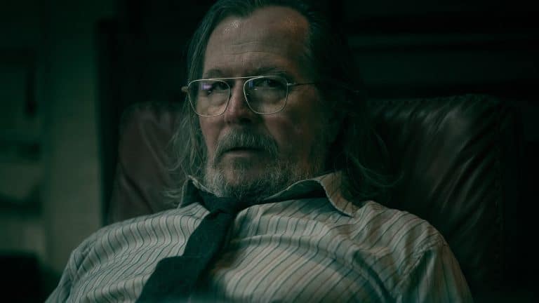 A middle-aged man with long gray hair, glasses, and a beard sits in a leather chair, wearing a striped shirt and a loosely knotted tie, evoking Gary Oldman's serious look from Slow Horses in the moody dim lighting.
