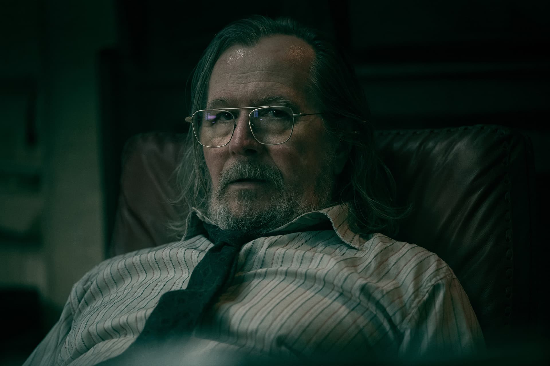 A middle-aged man with long gray hair, glasses, and a beard sits in a leather chair, wearing a striped shirt and a loosely knotted tie, evoking Gary Oldman's serious look from Slow Horses in the moody dim lighting.