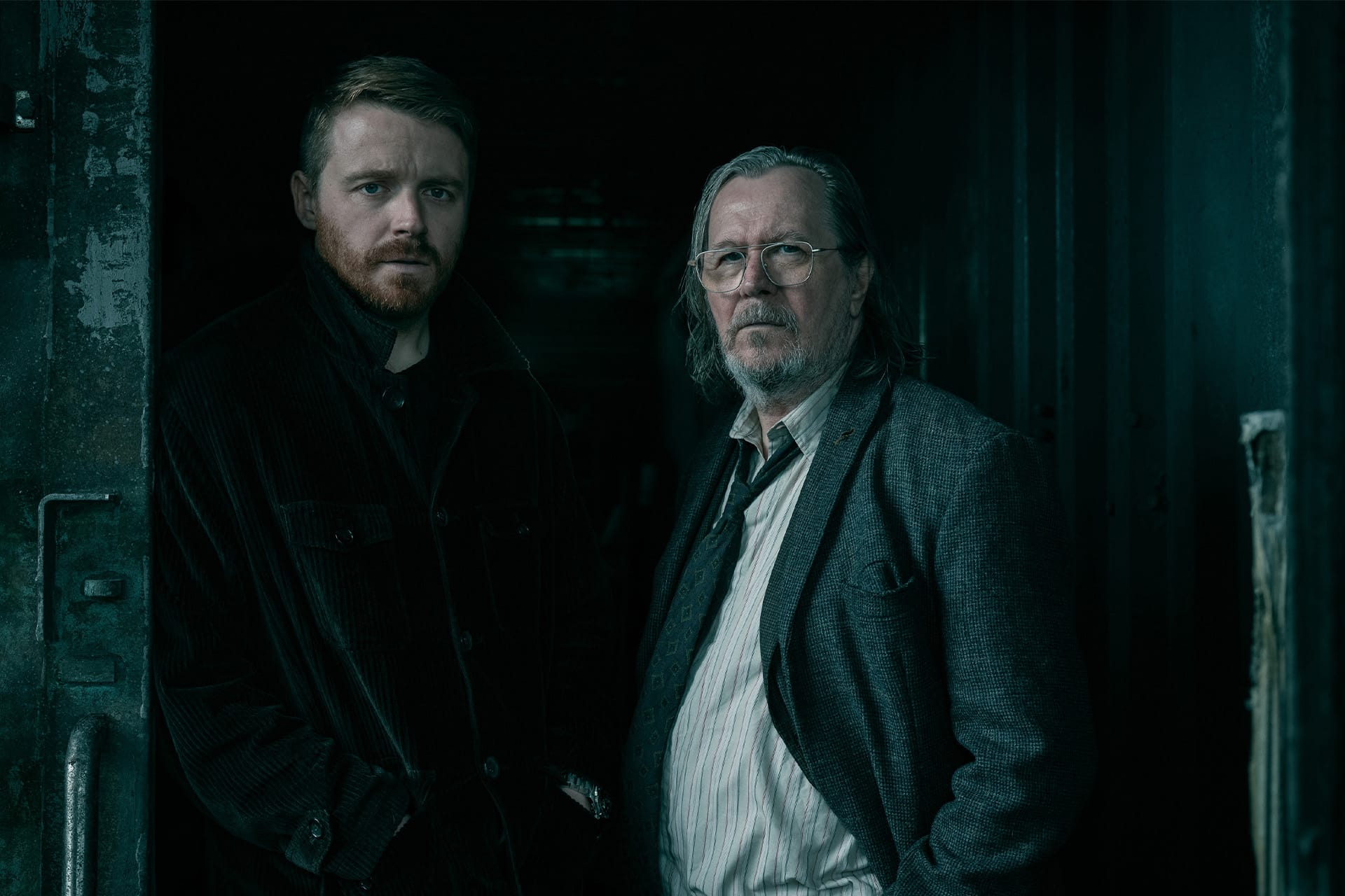 Two men stand side by side in a dimly lit, industrial setting. With serious expressions, they evoke the tense atmosphere of Slow Horses on Apple TV—one bearded with reddish hair in a dark coat, the other gray-haired in glasses and a striped shirt.