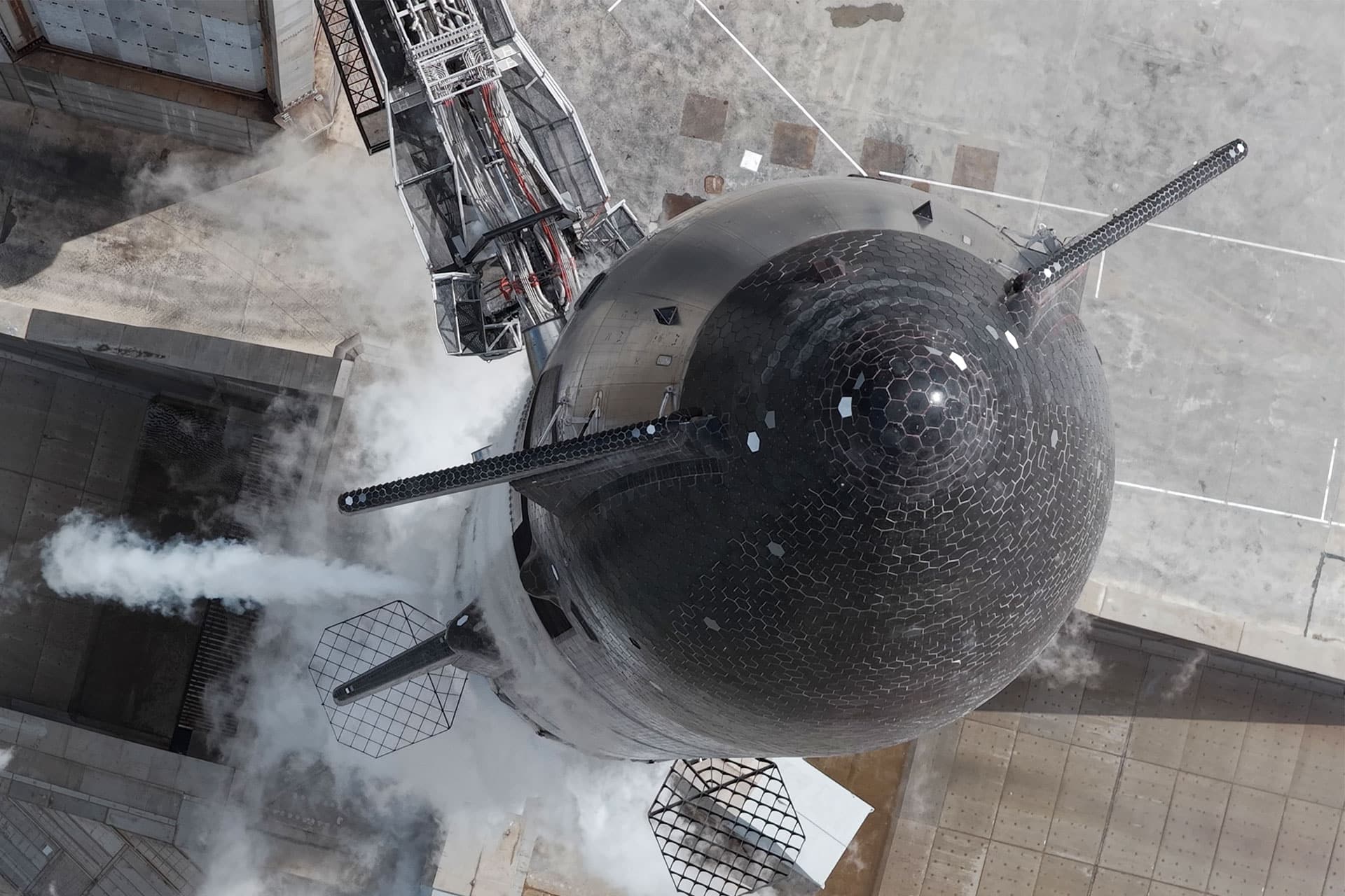 Aerial view of a large black SpaceX rocket on a launch pad releasing white vapor, with mechanical arms attached and hexagonal heat tiles visible on the rocket's surface. The concrete surroundings contrast with this unique part of the space ecosystem.