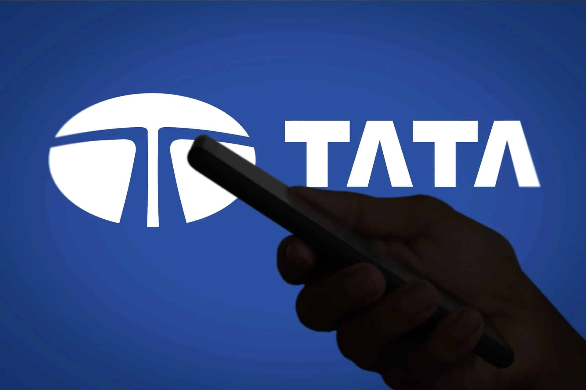 Tata Hosur - A hand holding a smartphone is silhouetted against a blue background featuring the Tata logo and the word "TATA" in bold white letters, highlighting Tata's growing role in India's iPhone supply chain.