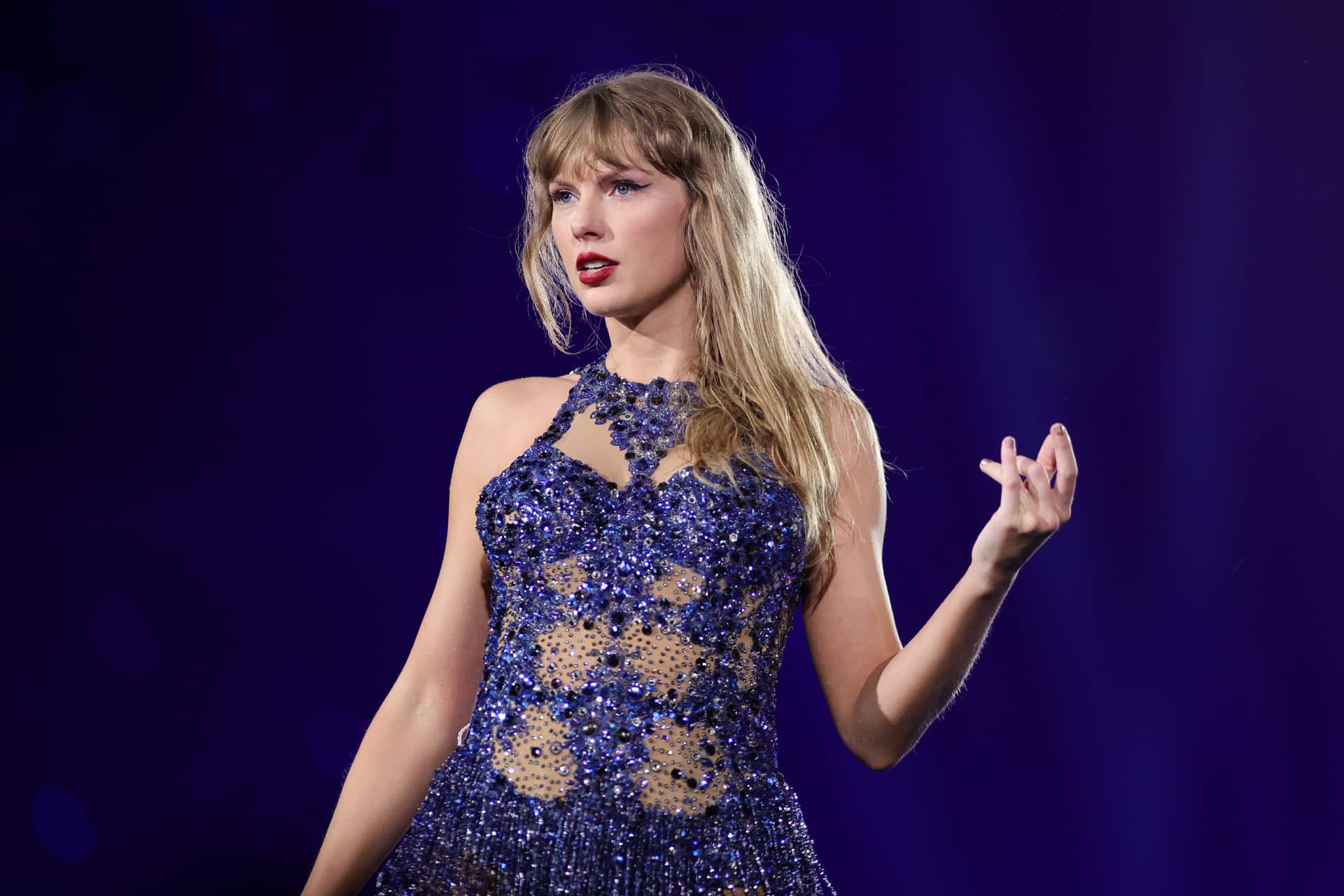 A woman with long blonde hair, reminiscent of Taylor Swift, wears a sparkling dark blue and beige dress. She stands on stage under dramatic purple lighting, looking to the side with one hand raised as if welcoming the future.