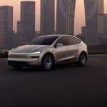 A sleek, modern white Tesla electric SUV drives on an empty highway at dusk, with tall glass skyscrapers and a hazy sky in the background. CarPlay route sharing enhances the connected driving experience.