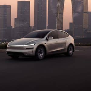 A sleek, modern white Tesla electric SUV drives on an empty highway at dusk, with tall glass skyscrapers and a hazy sky in the background. CarPlay route sharing enhances the connected driving experience.