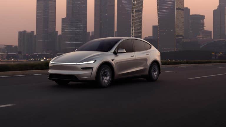 A sleek, modern white Tesla electric SUV drives on an empty highway at dusk, with tall glass skyscrapers and a hazy sky in the background. CarPlay route sharing enhances the connected driving experience.