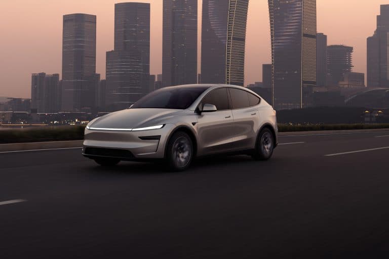 A sleek, modern white Tesla electric SUV drives on an empty highway at dusk, with tall glass skyscrapers and a hazy sky in the background. CarPlay route sharing enhances the connected driving experience.