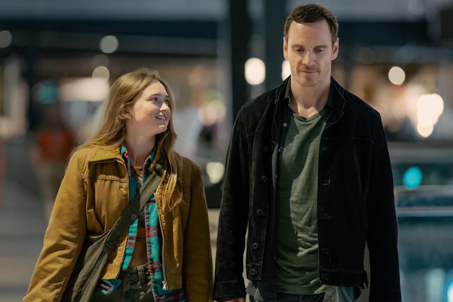 A young woman and a man walk indoors together. The woman, in a brown jacket and colorful scarf, smiles up at the man, who wears a dark jacket and green shirt. They appear to be having a friendly conversation, perhaps about their favorite Apple TV Originals.
