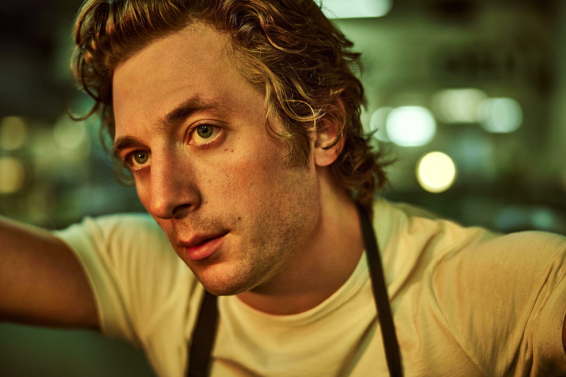 A young man with wavy blond hair, wearing a white t-shirt and dark apron, looks thoughtfully to the side in a warmly lit indoor setting, as if pondering what to watch next on streaming apps like Apple TV Originals.