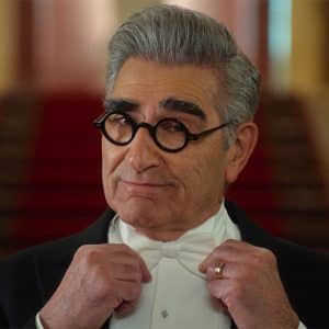 A man with gray hair and thick eyebrows, wearing round black glasses and a tuxedo, adjusts his bow tie while looking confidently at the camera, echoing the style seen in Schitt’s Creek. A red carpeted staircase is blurred in the background.
