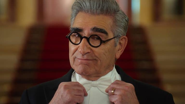 A man with gray hair and thick eyebrows, wearing round black glasses and a tuxedo, adjusts his bow tie while looking confidently at the camera, echoing the style seen in Schitt’s Creek. A red carpeted staircase is blurred in the background.