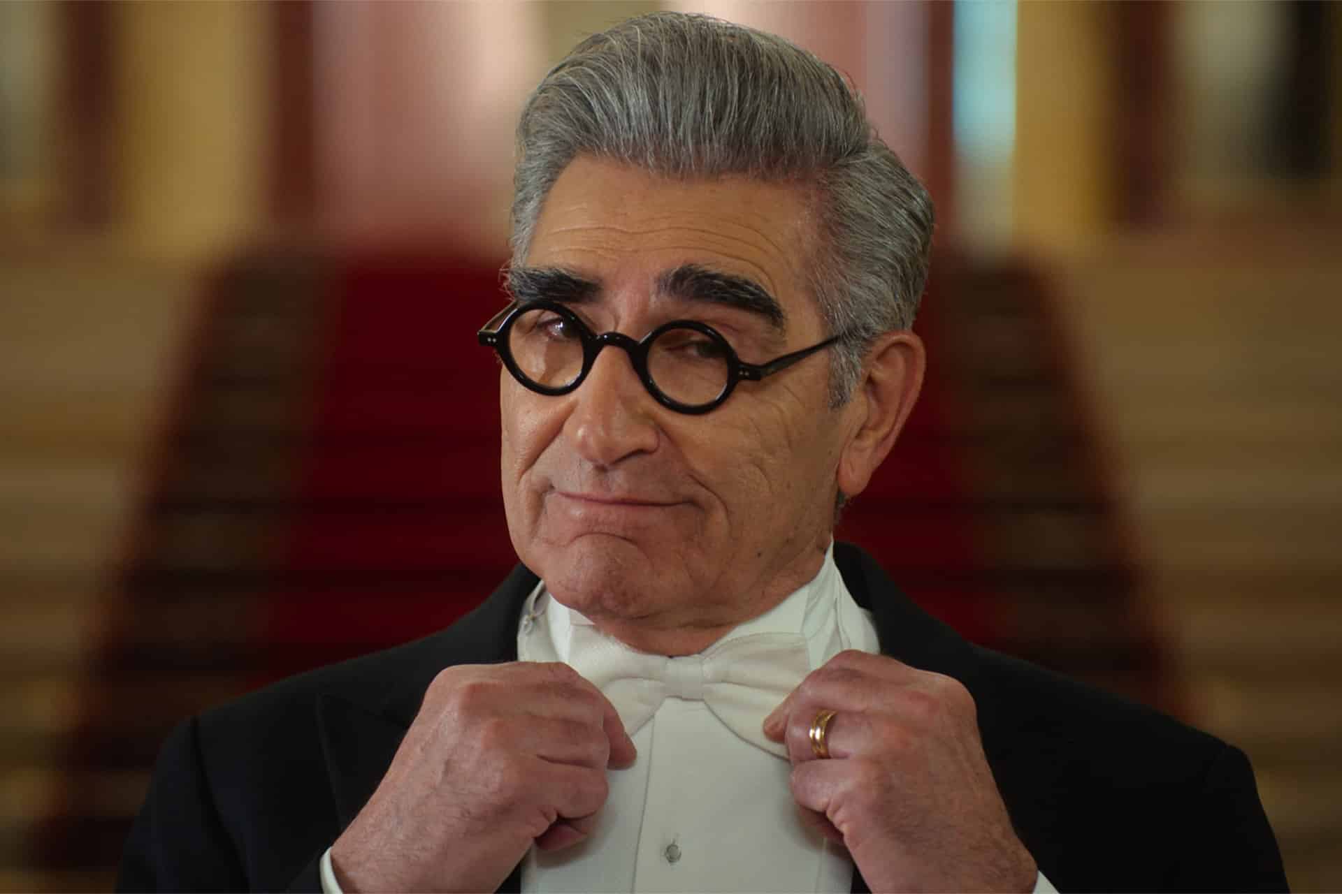 A man with gray hair and thick eyebrows, wearing round black glasses and a tuxedo, adjusts his bow tie while looking confidently at the camera, echoing the style seen in Schitt’s Creek. A red carpeted staircase is blurred in the background.