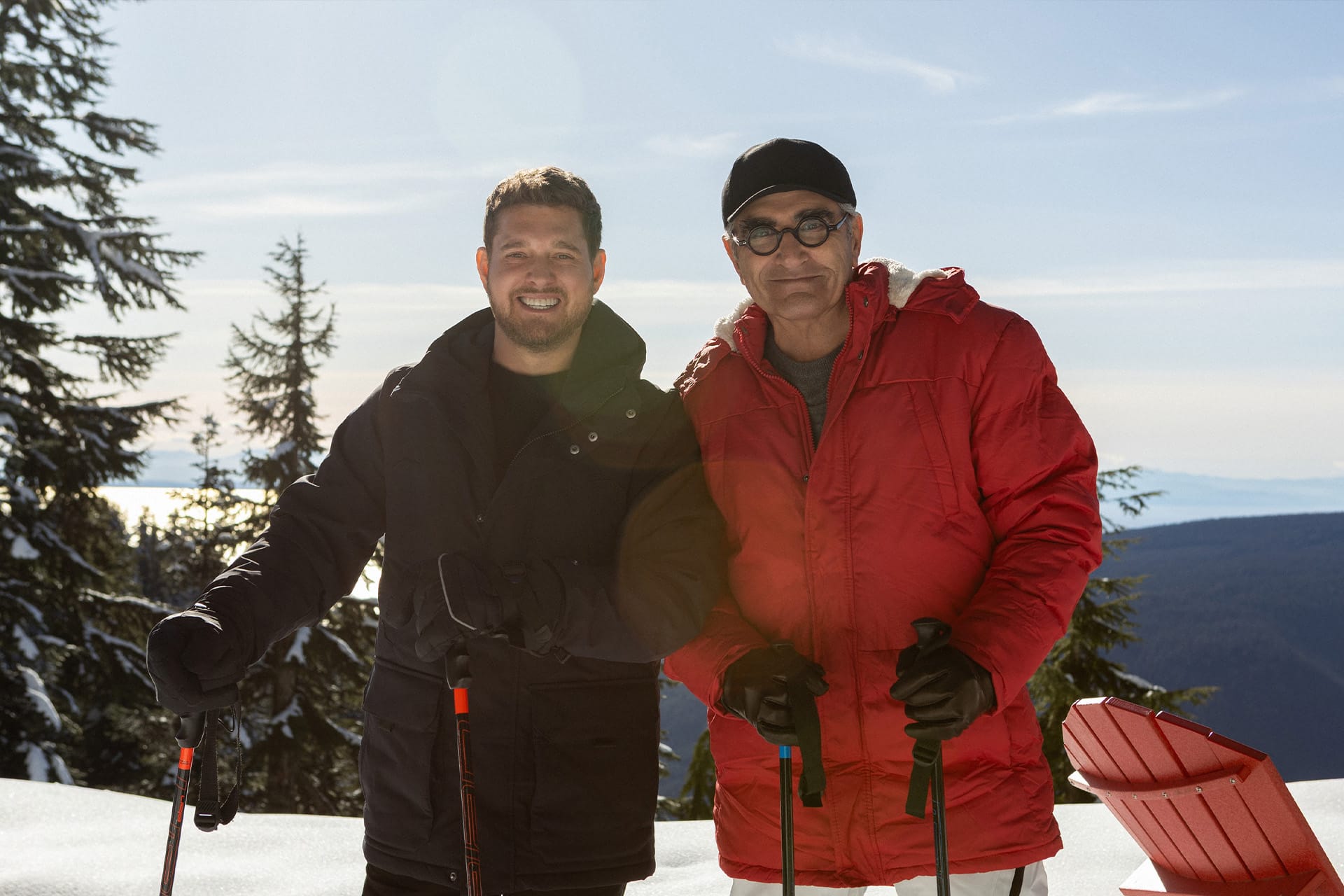 Two men dressed in winter jackets and gloves stand together on a snowy mountain, holding ski poles. With pine trees and a clear sky behind them, they smile at the camera—channeling an adventure worthy of The Reluctant Traveler with Eugene Levy.