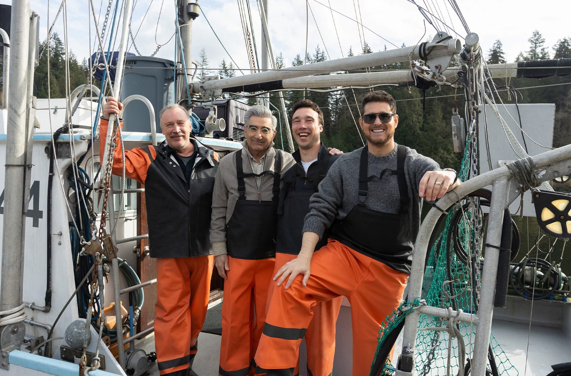 Four men in work overalls and orange waterproof pants stand smiling together on a fishing boat, surrounded by fishing gear, with a forested shoreline beyond—reminiscent of an adventure from an Apple Originals travel series.