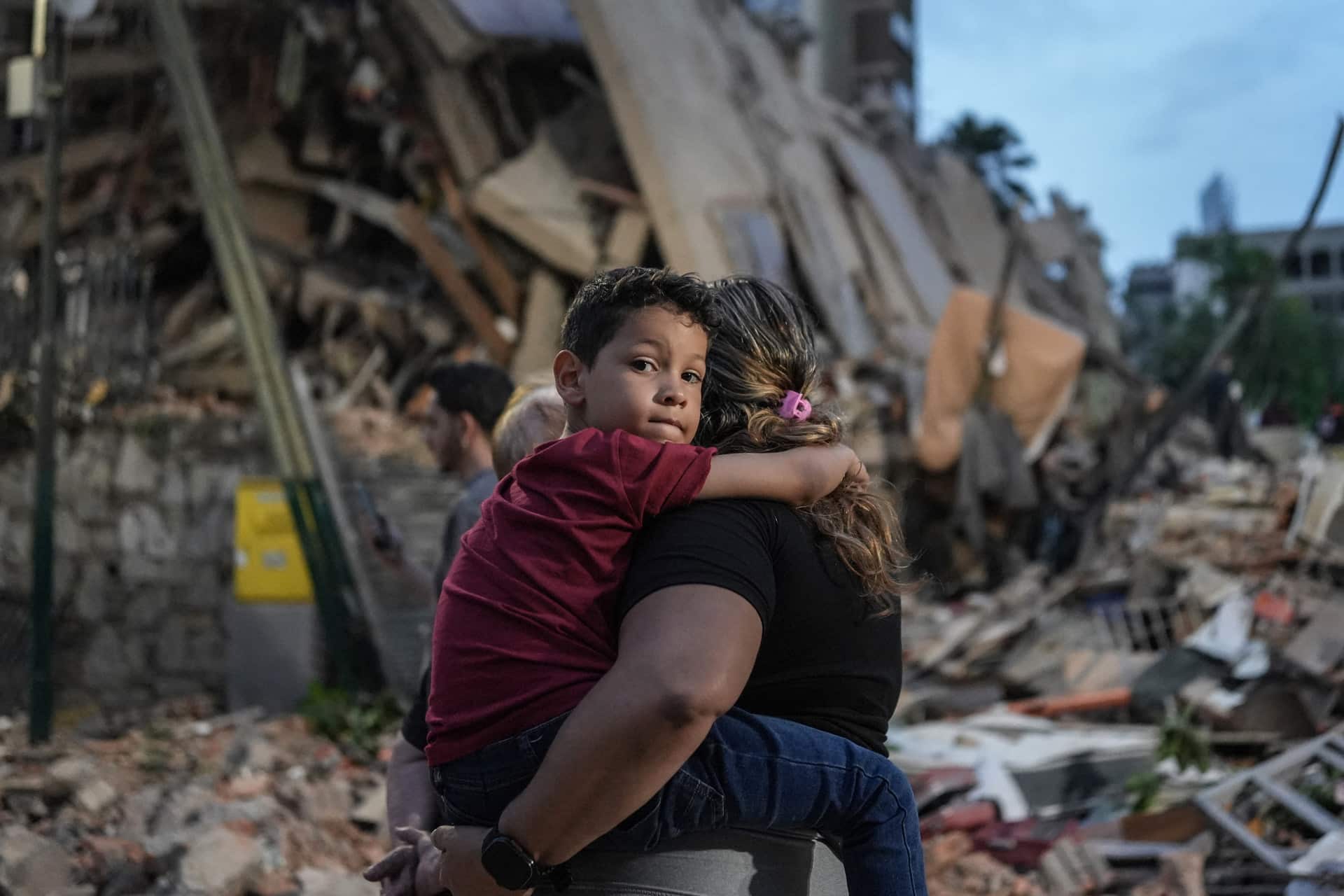 A woman carries a young boy through rubble in Venezuela, with a collapsed building in the background. The boy looks over her shoulder, appearing somber as they walk away from the destruction, highlighting the urgent need for disaster relief.