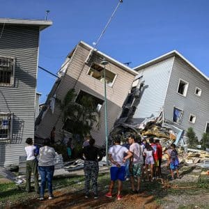 A group of people stand outside, looking at two multi-story buildings that have partially collapsed and are leaning against each other. Debris and rubble are scattered around as Apple Venezuela relief teams coordinate disaster relief under a clear blue sky.