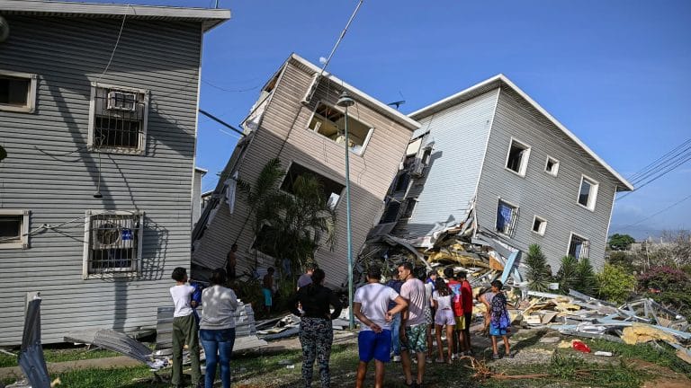 A group of people stand outside, looking at two multi-story buildings that have partially collapsed and are leaning against each other. Debris and rubble are scattered around as Apple Venezuela relief teams coordinate disaster relief under a clear blue sky.