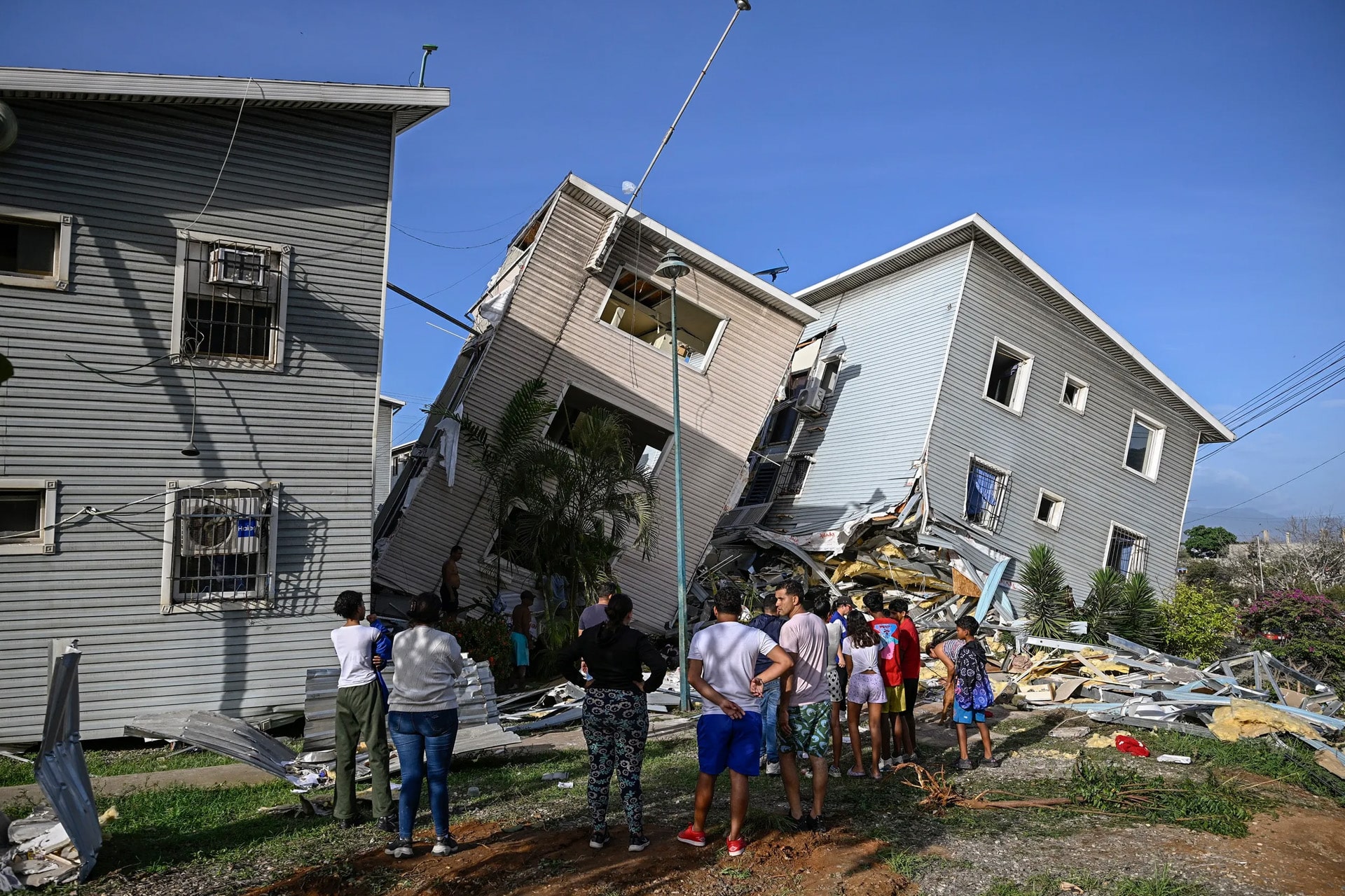 A group of people stand outside, looking at two multi-story buildings that have partially collapsed and are leaning against each other. Debris and rubble are scattered around as Apple Venezuela relief teams coordinate disaster relief under a clear blue sky.