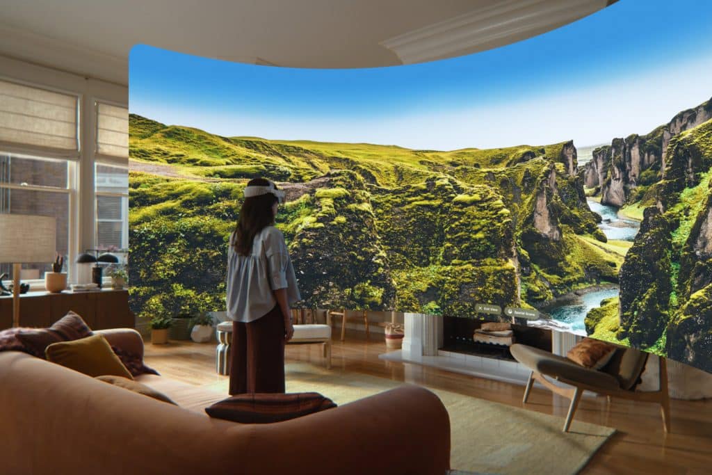 A person wearing a VR headset stands in a living room, using visionOS 27 to view an immersive virtual display of a lush, green canyon landscape that appears to surround them, blending nature with indoor space.