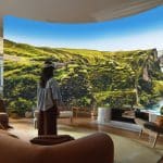 A person wearing a VR headset stands in a living room, using visionOS 27 to view an immersive virtual display of a lush, green canyon landscape that appears to surround them, blending nature with indoor space.