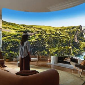 A person wearing a VR headset stands in a living room, using visionOS 27 to view an immersive virtual display of a lush, green canyon landscape that appears to surround them, blending nature with indoor space.