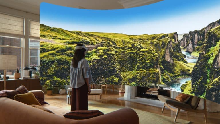 A person wearing a VR headset stands in a living room, using visionOS 27 to view an immersive virtual display of a lush, green canyon landscape that appears to surround them, blending nature with indoor space.