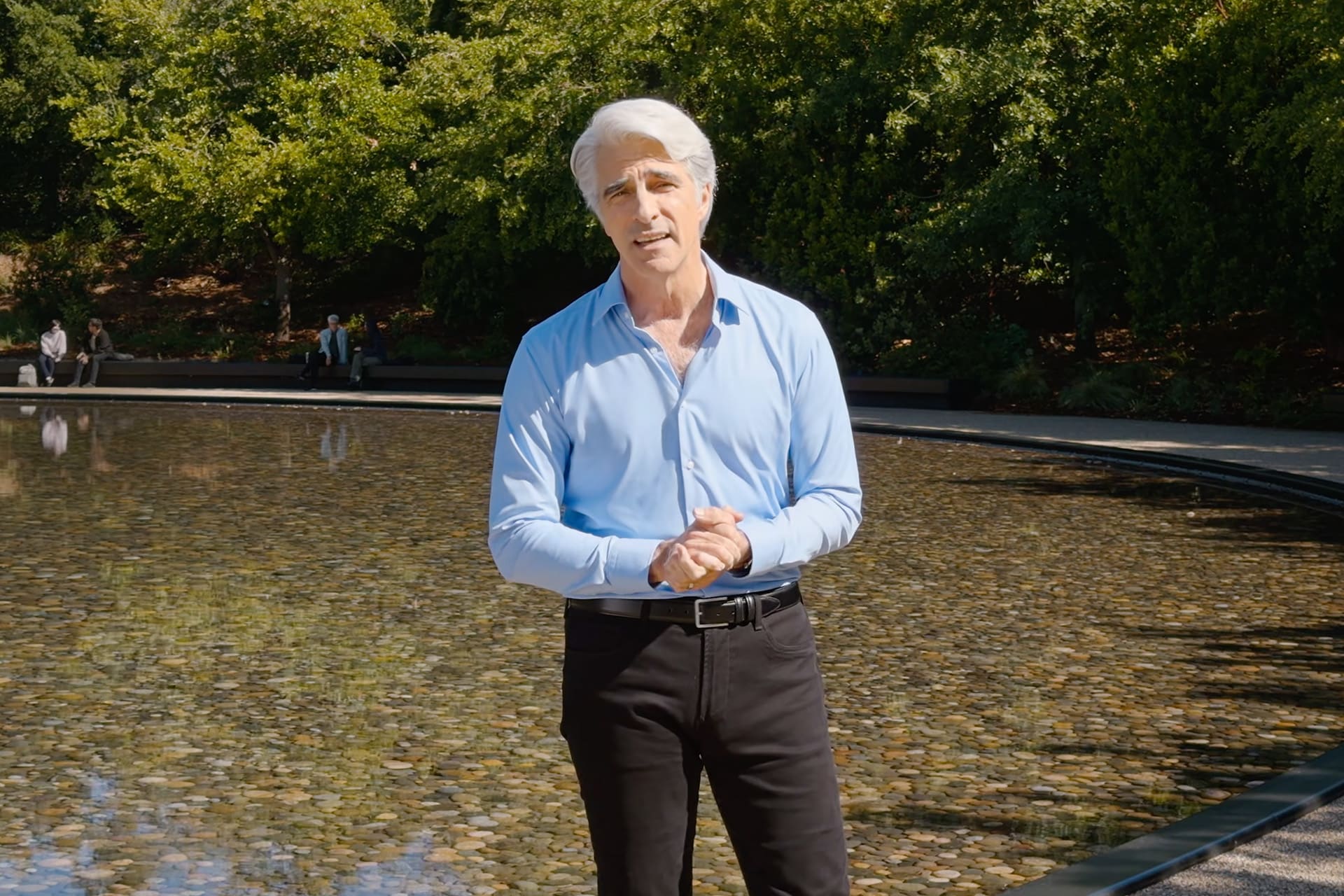 A man with gray hair wearing a light blue shirt and black pants stands in front of a shallow, circular reflecting pool, as trees and people sit in the background on a sunny day—reminiscent of an Apple keynote atmosphere.