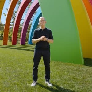 A man in a black shirt and glasses stands on green grass in front of large, colorful rainbow-shaped arches outdoors, capturing the moment with his iPhone 18 during WWDC26.