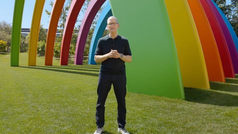 A man in a black shirt and glasses stands on green grass in front of large, colorful rainbow-shaped arches outdoors, capturing the moment with his iPhone 18 during WWDC26.