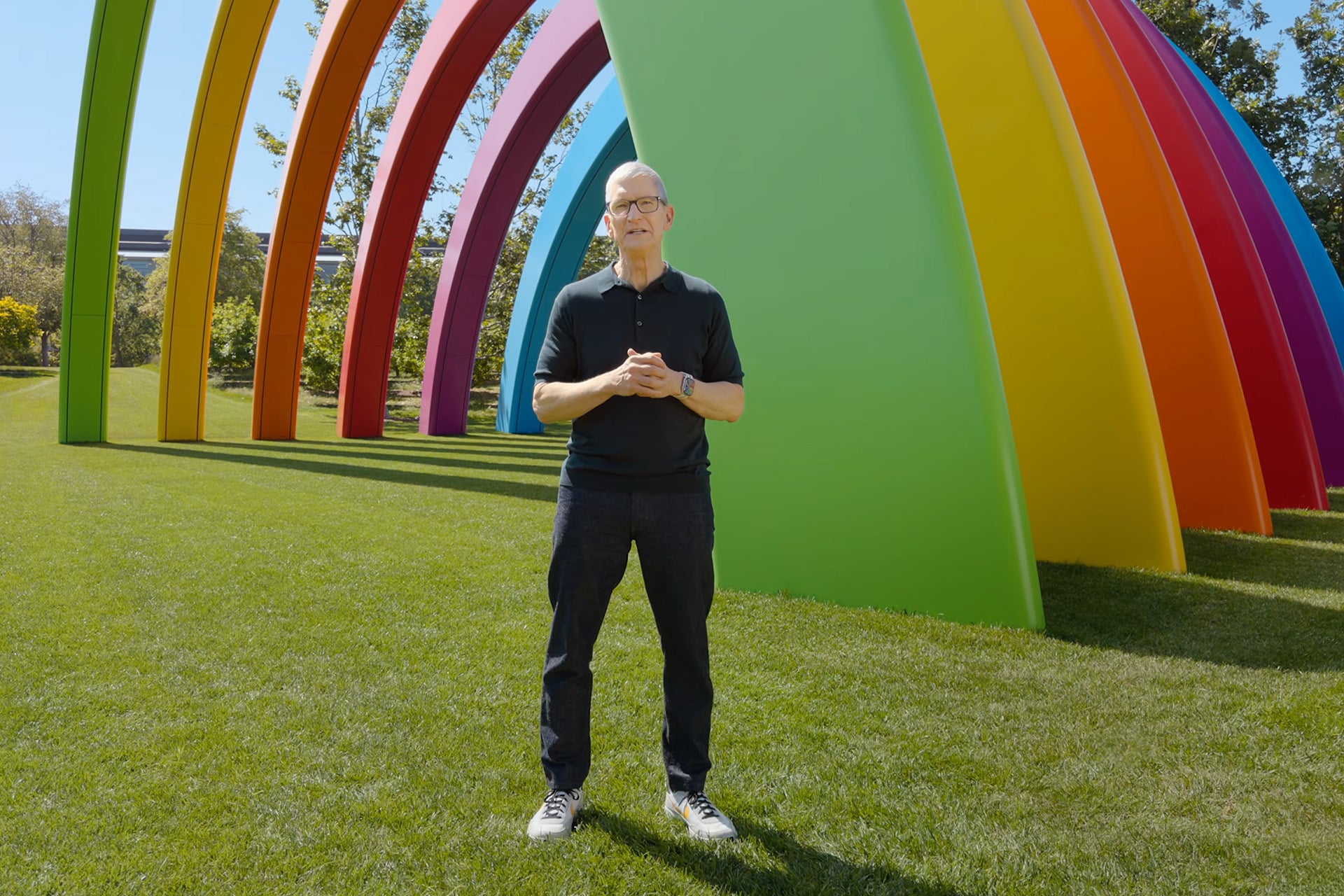A man in a black shirt and glasses stands on green grass in front of large, colorful rainbow-shaped arches outdoors, capturing the moment with his iPhone 18 during WWDC26.