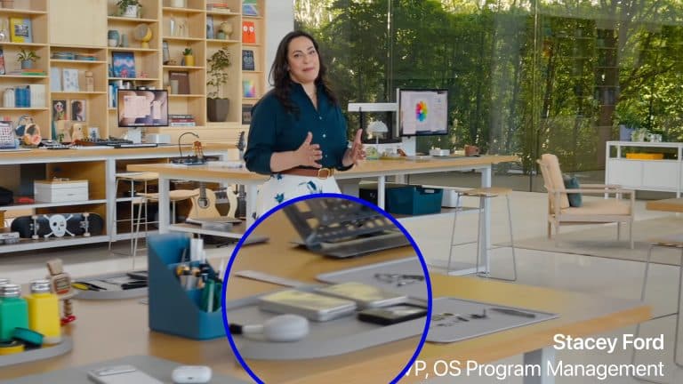 A woman stands in a modern office with large windows and shelves behind her. In the foreground, a table displays tech devices, including an iPad Fold highlighted with a blue circle. Text reads "Stacey Ford, VP, OS Program Management.