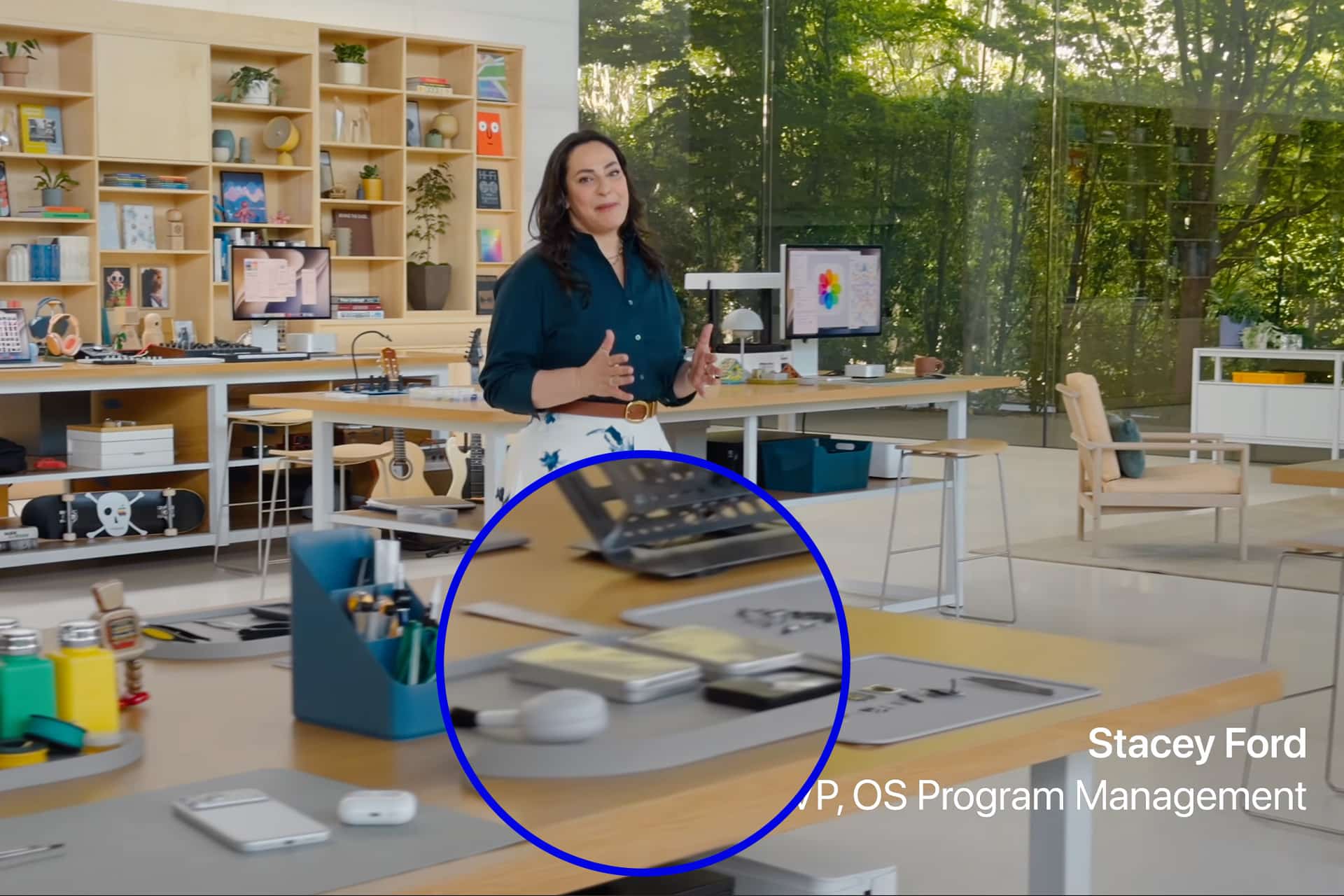 A woman stands in a modern office with large windows and shelves behind her. In the foreground, a table displays tech devices, including an iPad Fold highlighted with a blue circle. Text reads "Stacey Ford, VP, OS Program Management.