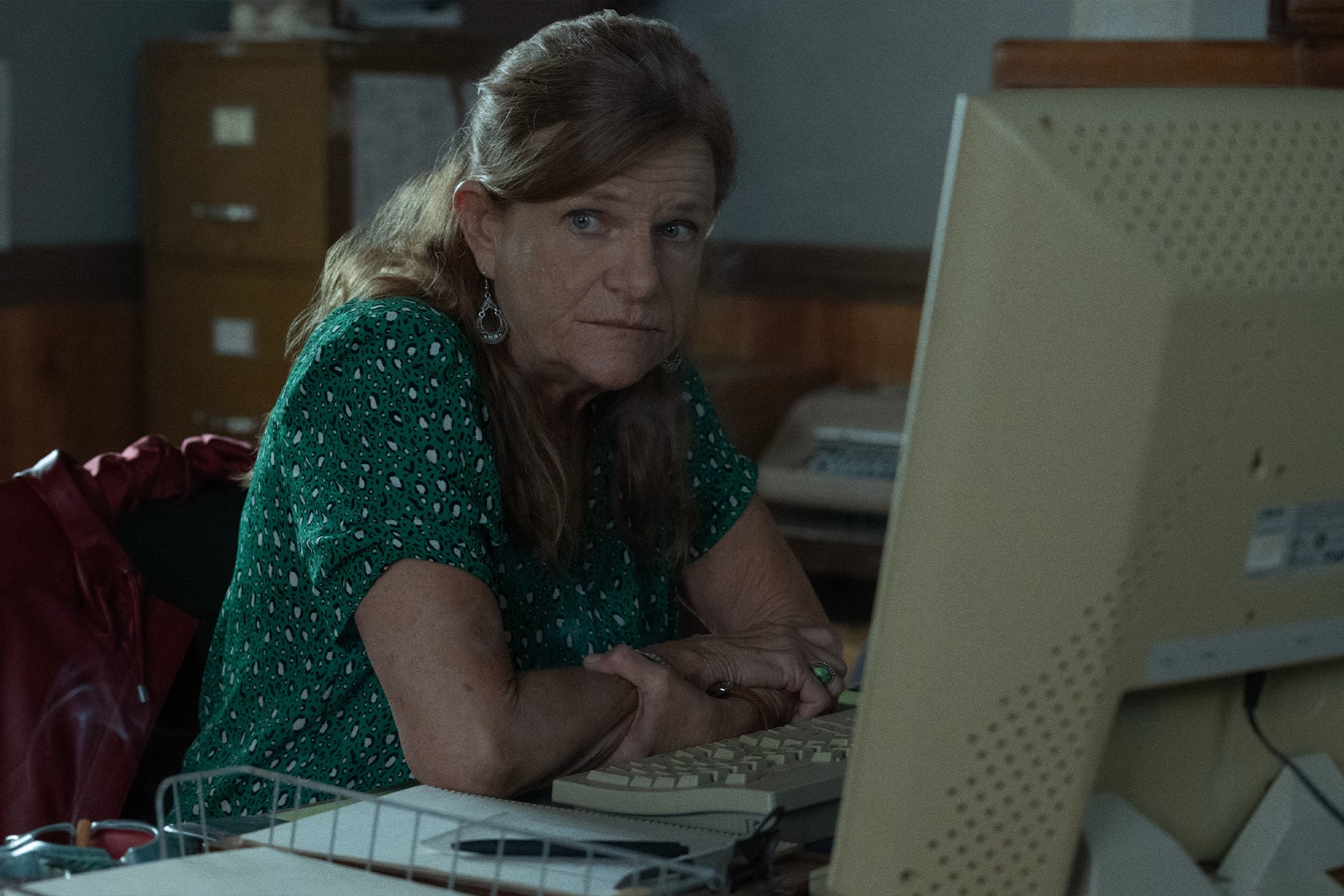 A woman with long brown hair wearing a green patterned shirt sits at a desk, looking seriously at her computer screen—perhaps searching for Easter eggs in an Apple TV drama—in an office with wooden walls and a filing cabinet behind her.