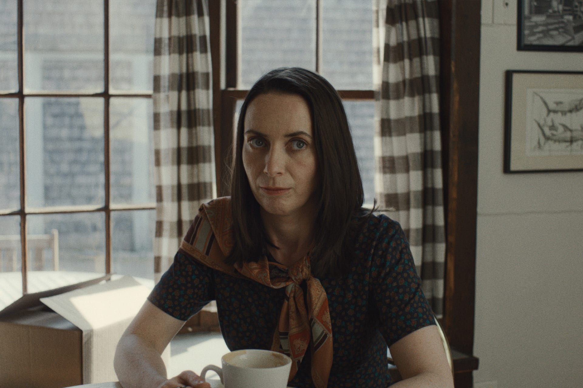A woman with straight dark hair sits at a table holding a white mug, looking at the camera. Behind her are large windows with striped curtains and framed artwork, creating a cozy scene reminiscent of a Widow’s Bay horror comedy on Apple TV.