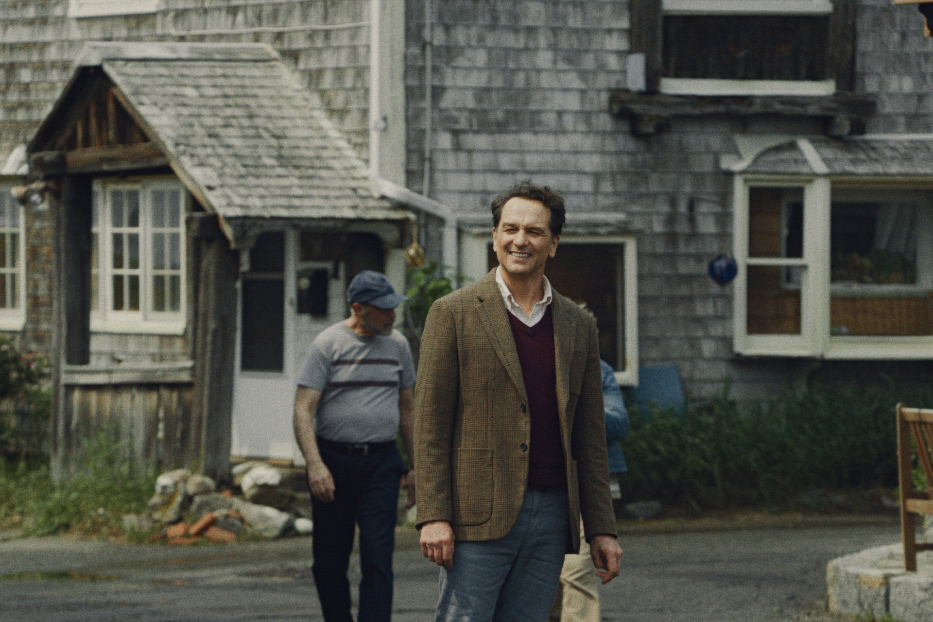 A man in a brown blazer and maroon sweater smiles while standing outside near a rustic wooden house in Widow’s Bay. Another person in a cap, partially obscured by the building, adds an air of mystery—like hidden Easter eggs on Apple TV.