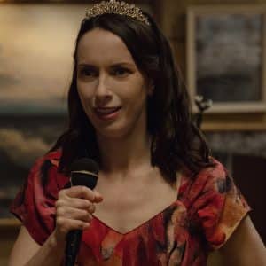 A woman with long brown hair wearing a tiara and a red patterned dress holds a microphone and smiles, standing indoors with framed art and warm lighting in the background—like a scene straight from Apple TV’s Widow’s Bay.
