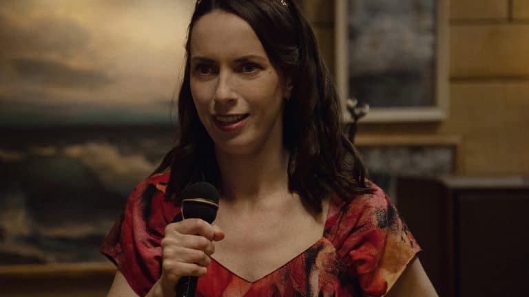 A woman with long brown hair wearing a tiara and a red patterned dress holds a microphone and smiles, standing indoors with framed art and warm lighting in the background—like a scene straight from Apple TV’s Widow’s Bay.