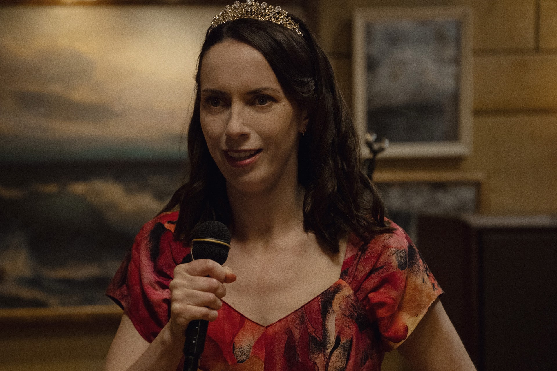 A woman with long brown hair wearing a tiara and a red patterned dress holds a microphone and smiles, standing indoors with framed art and warm lighting in the background—like a scene straight from Apple TV’s Widow’s Bay.