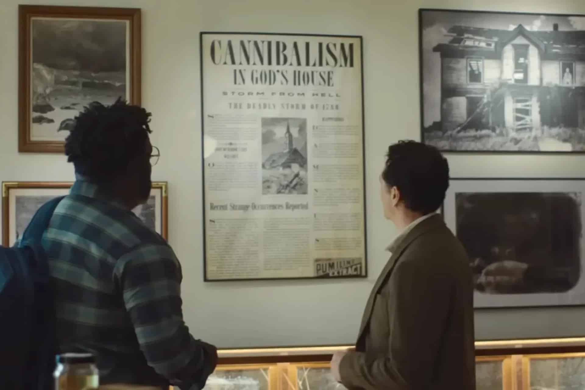 Two people stand in front of a wall display, examining a framed newspaper with the headline "Cannibalism in God's House." The wall features other black-and-white photographs, artwork, and subtle Easter eggs reminiscent of a horror comedy on Apple TV.