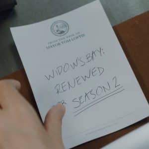 A hand holds a notepad labeled "From the desk of Mayor Tom Loftis" with a handwritten note that reads, "Widow's Bay season 2: Renewed on Apple TV," underlined for emphasis.