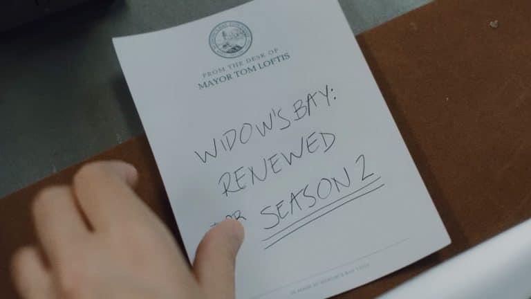 A hand holds a notepad labeled "From the desk of Mayor Tom Loftis" with a handwritten note that reads, "Widow's Bay season 2: Renewed on Apple TV," underlined for emphasis.