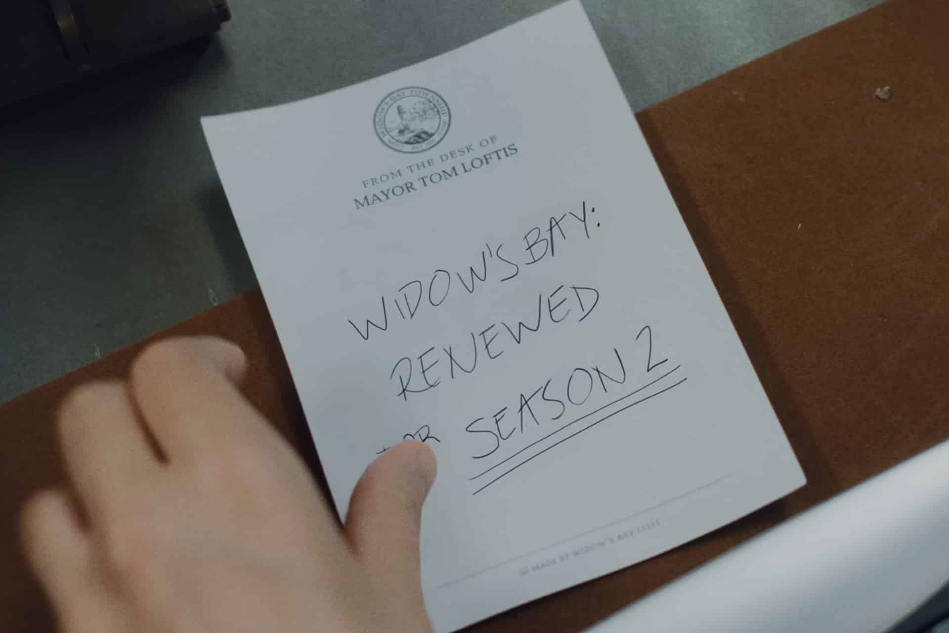 A hand holds a notepad labeled "From the desk of Mayor Tom Loftis" with a handwritten note that reads, "Widow's Bay season 2: Renewed on Apple TV," underlined for emphasis.